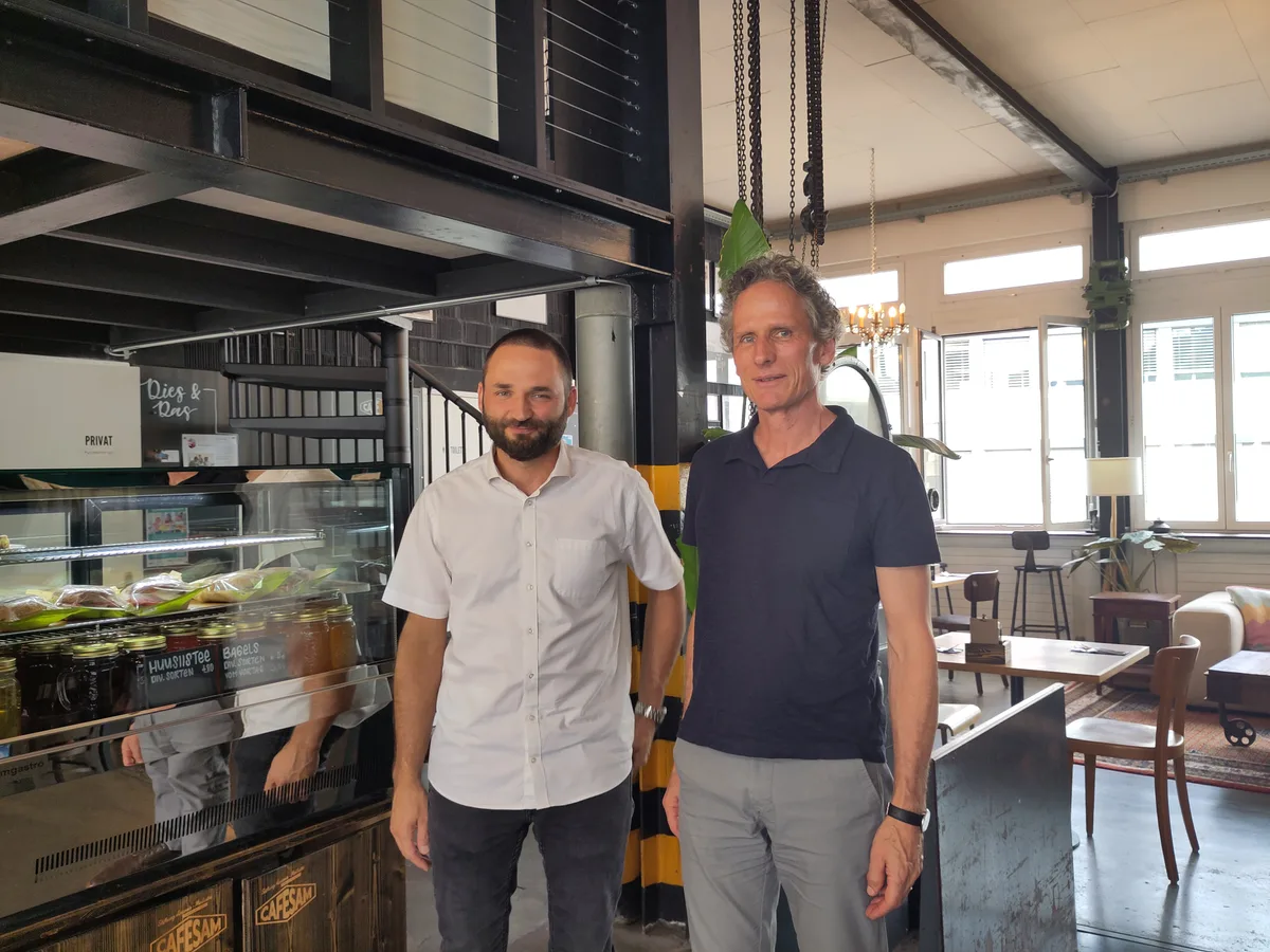 Dave Siegenthaler (links) und Leo Nissen wollen für alle Mitarbeitenden die passende Lösung finden. Man sieht zwei Männer vor der Vitrine eines Cafés.
